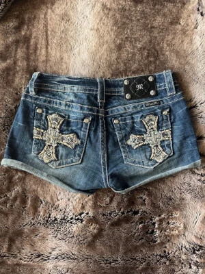 Blå denimshorts från Miss Me - Snygga blå jeansshorts från Miss Me med broderade kors och nitar på bakfickorna. Shorts med tydliga kontrastsömmar och coola detaljer som metallplatta med logga bak. 