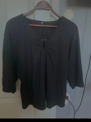 Svart topp med ringdetalj - Svart topp med v-ringning och snygg ringdetalj framtill. Trekvartsärm och lös passform gör den enkel att styla till olika looks. Perfekt för dig som gillar minimalistisk stil.
