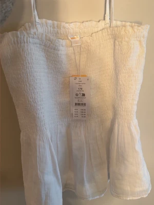 Vit smockad linnetopp med volang - Supersöt vit linnetopp från gina tricot i storlek 170 med smockad överdel och volang nertill. Toppen har tunna justerbara axelband och är tillverkad i ett lätt och luftigt material, perfekt för varma dagar. Helt oanvänd me prislapp! 