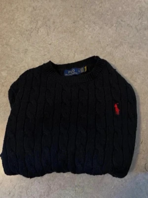 Mörkblå kabelstickad tröja Polo Ralph Lauren - Kabelstickad mörkblå tröja från Polo Ralph Lauren med klassisk röd logga broderad på bröstet. Tröjan har rund halsringning, långa ärmar och ribbade muddar. Perfekt för dig som gillar tidlös och stilren design.
