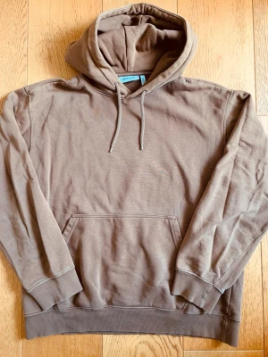 Brun hoodie från Weekday - Säljer en stilren brun hoodie från Weekday med klassisk känguruficka och dragsko i huvan. Hoodien är tillverkad i mjuk bomullsblandning och har ribbade muddar vid ärmslut och nederkant. Perfekt för en avslappnad och trendig look.