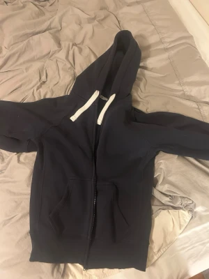 Mörkblå zip hoodie med vita snören. Russemerch - En stilren mörkblå hoodie med dragkedja och vita snören i huvan. Hoodien har känguruficka framtill och ribbade muddar vid ärmar och nederkant. Perfekt för en avslappnad look och enkel att matcha med jeans eller joggers.
