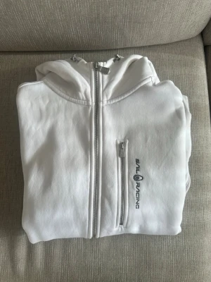 Vit Sail Racing hoodie med dragkedja herr - Vit hoodie från Sail Racing med hel dragkedja och huva. Modellen har en bröstficka med dragkedja och logotyp på framsidan. Materialet är mjukt och bekvämt, perfekt för en avslappnad stil. Snygg passform och klassisk design.