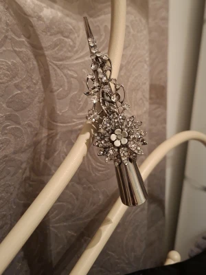 Silverfärgad hårklämma med strassblommor - Snygg hårklämma i silverfärgad metall med glittrande strass och blommönster. Klämman har en elegant, avlång form och dekoreras av små och stora blommor i kristall. Perfekt för att ge frisyren extra bling och stil.