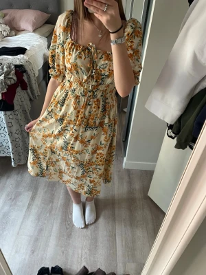 Gulblommig klänning med puffärm - Söt klänning med gul-orange blommönster på ljus botten. Modellen har puffärmar, smock i midjan och knytning vid bysten. Kjolen är lätt utställd och tyget är tunt och flowy, perfekt för vår och sommar. Sitter som S