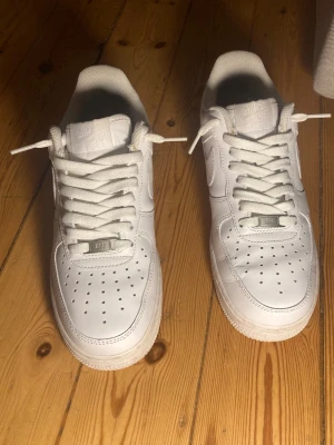 Nike Air Force 1 vita sneakers - Klassiska vita Nike Air Force 1 sneakers i lågt utförande. Skorna har en stilren design med perforerad tå, vita skosnören och Nike-logga på sidan. Tillverkade i läder med gummisula för bra grepp. Perfekta för en clean och tidlös look.