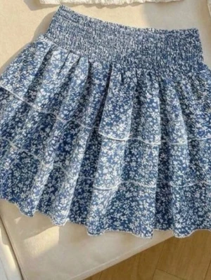 Blå blommig volangkjol - Supersöt blå kortkjol med vitt blommönster och volanger. Kjolen har bred smock i midjan för skön passform och är gjord i ett lätt, luftigt material som passar perfekt till varma dagar. Perfekt för dig som gillar en romantisk och trendig stil.