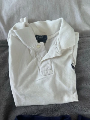 Vit pikétröja från Polo Ralph Lauren - S i barnstorlek! Klassisk vit pikétröja från Polo Ralph Lauren i 100% bomull. Tröjan har krage, knappar framtill och den ikoniska broderade loggan på bröstet. Perfekt för en clean och stilren look.