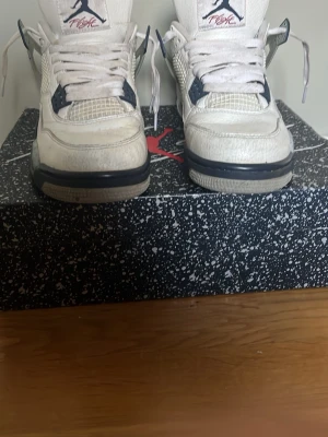 Air Jordan 4 Retro Vit/Grå/Blå - Snygga Air Jordan 4 Retro sneakers i vitt läder med grå och marinblå detaljer. Klassisk siluett med meshpaneler, synlig Air-enhet i sulan och Jumpman-logga på plösen. Perfekta för dig som gillar streetwear och ikoniska basketskor.