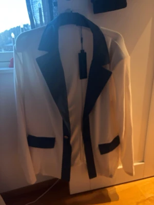 Vit kavaj/skjorta med svarta detaljer från COOFANDY - Stilren vit kavaj med svarta slag och svarta detaljer vid fickorna. Tunnt och luftigt material perfekt till sommaren med klassisk passform samt lång ärm och enkelknäppning framtill. Perfekt för en trendig och uppklädd look.