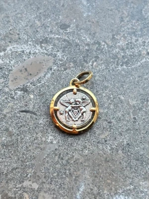 Rund berlock Amor Fati i metall - Unik rund berlock i silver- och guldfärgad metall med detaljerat motiv av två figurer och hjärta på framsidan. Baksidan har texten 'AMOR FATI'. Perfekt som personlig detalj till halsband eller armband.