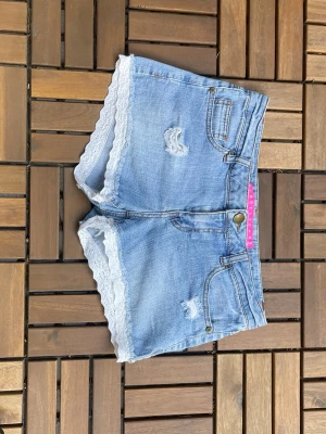 Ljusa jeansshorts med spetskant - Snygga ljusblå jeansshorts med slitna detaljer och vit spetskant längst ner. Klassisk femficksmodell med knapp och dragkedja framtill. Perfekta för en avslappnad och trendig look i sommar. Midjemått 39.5-42cm❣️