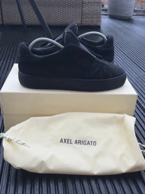 Svrata Axel Arigato laceless - Stilrena svarta sneakers från Axel Arigato i mocka med helsvart sula. Klassisk low-top modell med minimalistisk design och diskreta detaljer. Perfekta för dig som gillar en clean och modern look. Skorna levereras med originalpåse.