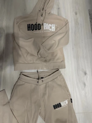Beige hoodie set med dragkedja från Hoodrich - Beige hoodie från Hoodrich med dragkedja framtill, huva med dragsko och stor tryckt logga på bröstet. Tillverkad i mjukt och fleeceliknande material för extra komfort. Perfekt för en avslappnad streetwear-look.