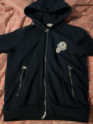  Moncler hoodie med dragkedja - Snygg svart hoodie från Moncler med hel dragkedja framtill, två fickor med dragkedja och klassisk Moncler-logga på bröstet. Hoodien har huva med dragsko och är tillverkad i mjukt material som känns skönt mot huden. Perfekt för en avslappnad och trendig stil.