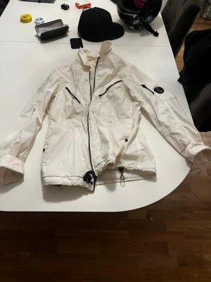 Vit vindjacka från cp company  - Snygg vit vindjacka fråm cp company dragkedjor och justerbar midja. Jackan har flera fickor med dragkedja, resår vid ärmslut och en cool detalj på ena ärmen. Perfekt för blåsiga dagar och enkel att matcha med olika outfits. Den är ÄKTA!!