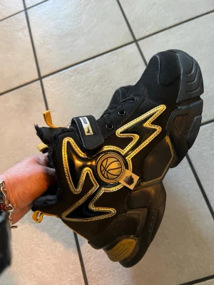 Svarta och guldiga sneakers med basketdetalj - Unika svarta sneakers  med coola guldiga detaljer och en basketboll-logga på sidan. Skorna har chunky sula, kardborreband och dragflikar i gult. Perfekta för dig som vill sticka ut med sportig stil.