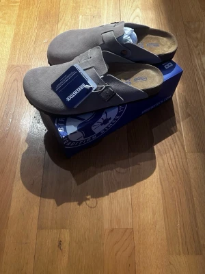 Birkenstock Boston tofflor grå mocka - Snygga grå/ beigea Birkenstock Boston tofflor i mocka med justerbart spänne och klassisk korksula. Allt ingår som visas i artikeln. Säljer då ja fick dessa i present och de var i för liten storlek! Bara att ställa frågor! 🙌🏼
