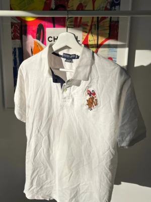 Vit pikétröja från U.S. Polo Assn. - Klassisk vit pikétröja från U.S. Polo Assn. med broderad logga på bröstet och mörkblå detaljer i kragen. Tillverkad i 100% bomull och har korta ärmar samt knappar vid halsen. Perfekt för en clean och sportig look. Storlek XL i junior, dvs M i senior. 