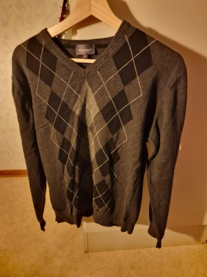 Grå kofta - Snygg grå tröja från Atlany med klassiskt argylemönster i svart och ljusgrått framtill. V-ringad modell med långa ärmar och ribbade muddar. Perfekt för en stilren och tidlös look.