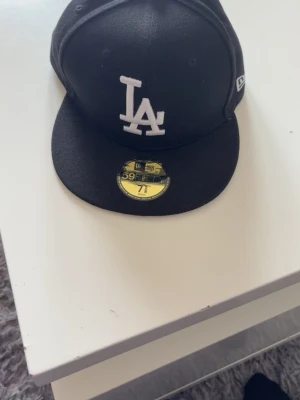 Svart LA-keps från New Era 59Fifty - Snygg svart keps från New Era 59Fifty med LA-logga broderad i vitt framtill. Klassisk flat peak och strukturerad passform. Perfekt för dig som gillar streetstyle och sportiga vibes.