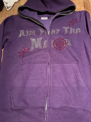 Lila hoodie med tryck och dragkedja - Lila hoodie med huva och dragkedja framtill. På bröstet står det 'Aim For The Moon' i glittrigt tryck och röda detaljer med stjärnmotiv. Två stora fickor framtill. Perfekt för en chill och avslappnad stil.