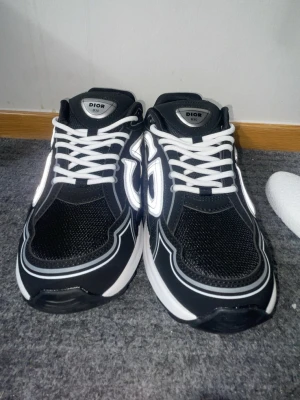 Svarta Dior B30 sneakers med vita detaljer - Snygga svarta Dior B30 sneakers med vita och reflekterande detaljer. Skorna har vit sula, vita skosnören och meshpaneler för extra andningsförmåga. Stora Dior-loggor på sidorna och modern chunky design. Perfekta för dig som gillar exklusiva och trendiga sneakers.