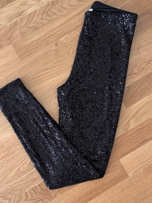 Svarta paljettleggings - Glittriga svarta leggings täckta med paljetter. De har en tight passform och resår i midjan för extra komfort. Perfekta för att sticka ut och skapa en cool look. Materialet är stretchigt och följsamt.