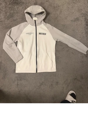 Vit och grå hoodie från Hoodrich - Snygg hoodie från Hoodrich i vitt med ljusgrå ärmar och huva. Dragkedja framtill, tryck med 'HOODRICH' på bröstet och stor text på ryggen. Perfekt för en streetwear-look. Tillverkad i mjukt material för skön känsla.