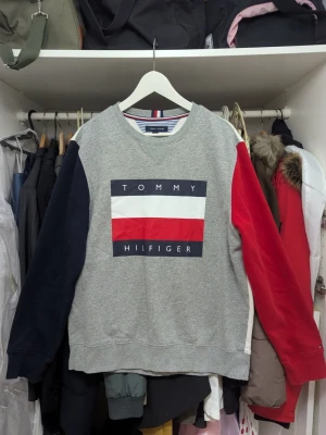 Multi-color långärmad tröja Tommy Hilfiger - Grå långärmad tröja från Tommy Hilfiger med stor logga framtill. Ena ärmen är marinblå och den andra röd, vilket ger en snygg kontrast. Tröjan har rund halsringning och är gjord i mjuk bomull. Perfekt för dig som gillar klassiska märken och streetstyle.