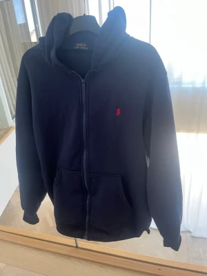 Mörkblå hoodie från Polo Ralph Lauren - Mörkblå hoodie med dragkedja från Polo Ralph Lauren. Klassisk design med huva, två fickor framtill och röd broderad logga på bröstet. Mjuk och skön bomullsblandning, perfekt för en avslappnad stil. 