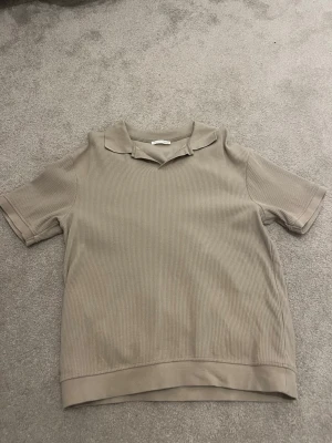 Beige ribbad polotopp från Zara - Säljer en oanvänd beige ribbad polotopp från Zara med korta ärmar och avslappnad krage. Toppen har en mjuk känsla och är perfekt för en chill och stilren look. Passar dig som gillar neutrala färger och enkel design.