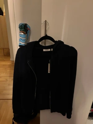 Mörkblå hoodie från RYVLS - Säljer en mörkblå stickad hoodie från RYVLS Stockholm. Modellen heter Justin Soft Knit Zip Hoodie och har dragkedja framtill samt huva. Perfekt för dig som gillar en stilren och enkel look. Materialet är mjukt och skönt mot huden.