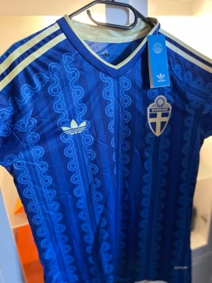 Blå Sverige fotbollströja Adidas - Säljer en blå Sverige fotbollströja från Adidas med gulddetaljer och mönstrad design. Tröjan har korta ärmar, V-ringning och klassiska Adidas-ränder på axlarna. Sverige-emblem på bröstet och Adidas-logga. Perfekt för dig som älskar fotboll och vill visa ditt stöd.