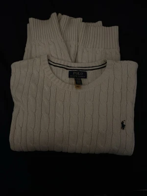 Vit kabelstickad tröja Polo Ralph Lauren - Kabelstickad vit tröja från Polo Ralph Lauren med rund halsringning och klassisk broderad logga på bröstet. Tröjan har ribbade muddar vid ärmslut och nederkant och är tillverkad i mjukt material som passar perfekt till säsongens outfits.