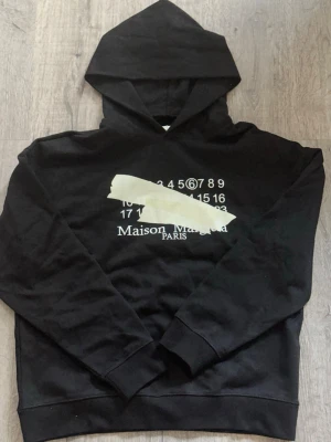 Maison Margiela svart hoodie - Svart hoodie från Maison Margiela med ikoniskt tryck framtill i vitt och beige. Klassisk huva och ribbade muddar. Oversized fit och tillverkad i mjuk bomull. Perfekt för en trendig och avslappnad stil.