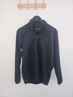 Svart träningströja från Under Armour - Snygg svart långärmad träningströja från Under Armour med halv dragkedja framtill och diskret logga på bröstet. Tillverkad i ett mjukt och stretchigt funktionsmaterial som passar perfekt för träning eller uppvärmning.