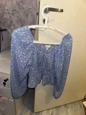 Blå blommig blus från H&M - Söt blå blus från H&M med vitt blommönster och långa ärmar. Blusen har en lös passform, resår i midjan och knytning framtill vid halsen. Perfekt för en trendig och avslappnad look. Materialet är lätt och luftigt, vilket gör den skön att bära.