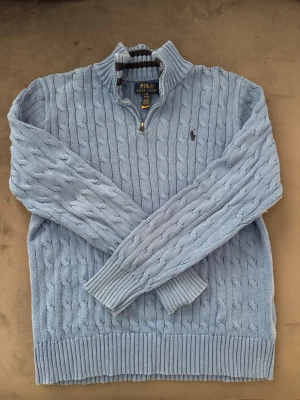 Ljusblå kabelstickad tröja Polo Ralph Lauren - Ljusblå kabelstickad tröja från Polo Ralph Lauren med halv dragkedja och hög krage. Tröjan har ribbade muddar och nederkant samt den klassiska broderade loggan på bröstet. Perfekt för dig som vill se stilig ut till allt.