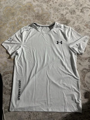 Vit träningströja från Under Armour - Snygg vit träningströja från Under Armour med svarta loggor och textdetaljer på bröstet, ärmen och längs sidan. Tillverkad i ett lätt och snabbtorkande funktionsmaterial, perfekt för intensiva träningspass. Rund halsringning och korta ärmar.