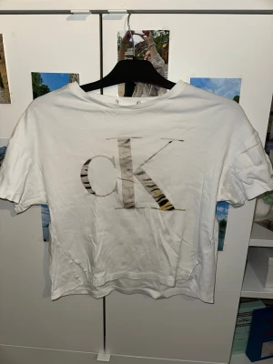 Vit Calvin Klein t-shirt med logga - Vit t-shirt från Calvin Klein Jeans med stor cK-logga i beige och svart tigermönster på bröstet. Klassisk passform med rund halsringning och korta ärmar. Tillverkad i mjukt bomullsmaterial. Perfekt för en stilren och trendig look.