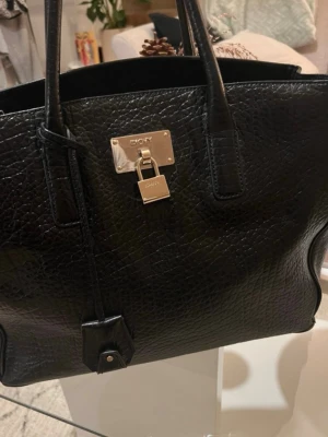 Svart handväska från DKNY - Snygg svart handväska från DKNY i präglat läder med guldfärgade metalldetaljer och hänglås framtill. Väskan har två rejäla handtag och en stilren, strukturerad form som ger en lyxig känsla. Perfekt för dig som gillar klassisk och trendig design.