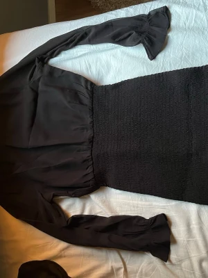 Svart långärmad klänning med smock - Stilren svart klänning med långärmade, lätt utsvängda ärmar och smockad midja samt kjol. Överdelen är i ett mjukt, flowy tyg som ger en snygg kontrast mot den åtsittande nederdelen. Perfekt för dig som gillar en enkel men trendig look.