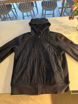 Svart cardigan från Polo Ralph Lauren - Snygg svart cardigan från Polo Ralph Lauren med dragkedja framtill och justerbar huva. Tröjan har det klassiska Polo-logotrycket på bröstet och är tillverkad i mjukt material. Perfekt för en avslappnad och stilren look.