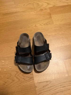 Svarta Birkenstock sandaler med spännen - Svarta sandaler från Birkenstock med två breda remmar och justerbara metallspännen. Klassisk korksula och anatomisk fotbädd för extra komfort. Perfekta för en avslappnad och trendig stil. Materialet är läder och kork.