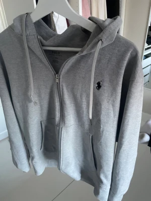 Grå zip hoodie från Polo Ralph Lauren - Snygg grå zip hoodie från Polo Ralph Lauren med klassisk huva och dragsko. Tröjan har fickor framtill och den ikoniska svarta loggan broderad på bröstet. Tillverkad i mjukt bomullsmaterial som är skönt att ha på sig. Perfekt för en chill och stilren look.