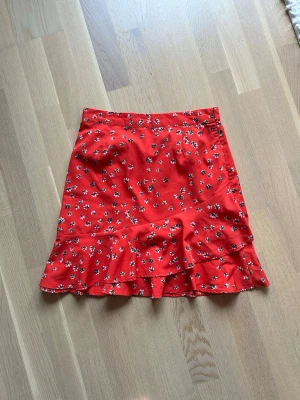Röd blommig volangkjol - Superfin röd kortkjol med små vita och svarta blommor och volang nertill. Perfekt för dig som vill ha en färgglad och söt kjol med lite extra detalj. Kjolen har en feminin passform och är lätt att matcha med både t-shirt och blus.