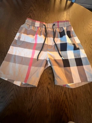 Shorts  - Snygga rutiga shorts med beige, svart, vit och röd färg. Shortsen har elastisk midja med svart dragsko och två sidofickor. Perfekta för sommardagar och har en klassisk design som sticker ut.