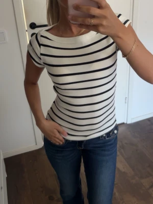 Randig vit t-shirt med svarta ränder - Snygg vit t-shirt med svarta horisontella ränder och rund hals. Modellen är figurnära med korta ärmar och tillverkad i ett mjukt, ribbat material som sitter skönt på kroppen. Perfekt till jeans eller kjol för en klassisk look.