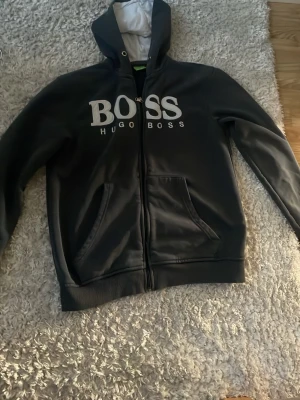 Svart hoodie från Hugo Boss - Svart hoodie från Hugo Boss med stor vit logga på framsidan. Dragkedja framtill och huva med snörning. Klassisk streetwear-stil, perfekt för dig som gillar snygga och bekväma plagg. Materialet känns mjukt och skönt mot huden.